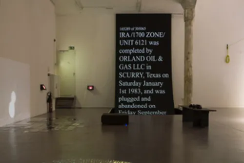 An impression of an installation: A room with white walls and an old column in the middle; there's a projection on the left, a beamer in the middle, and a bank on the right. On the background, a big black screen with the words »IRA / 1700 Zone / UNIT 6121 was completed by ORLAND OIL & GAS LLC in SCURRY, Texas on Saturday, January 1st, 1983, and was plugged and abandoned...«